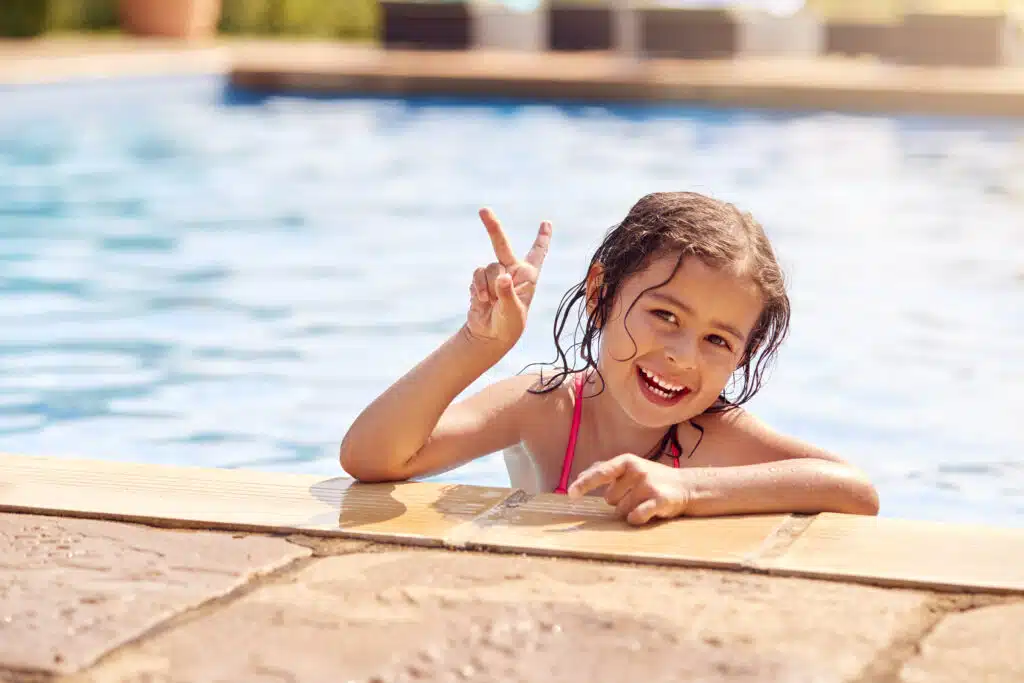 Portrait Of Young Girl Leaning On Edge Of Swimming Pool Making Peace Sign On Summer Vacation